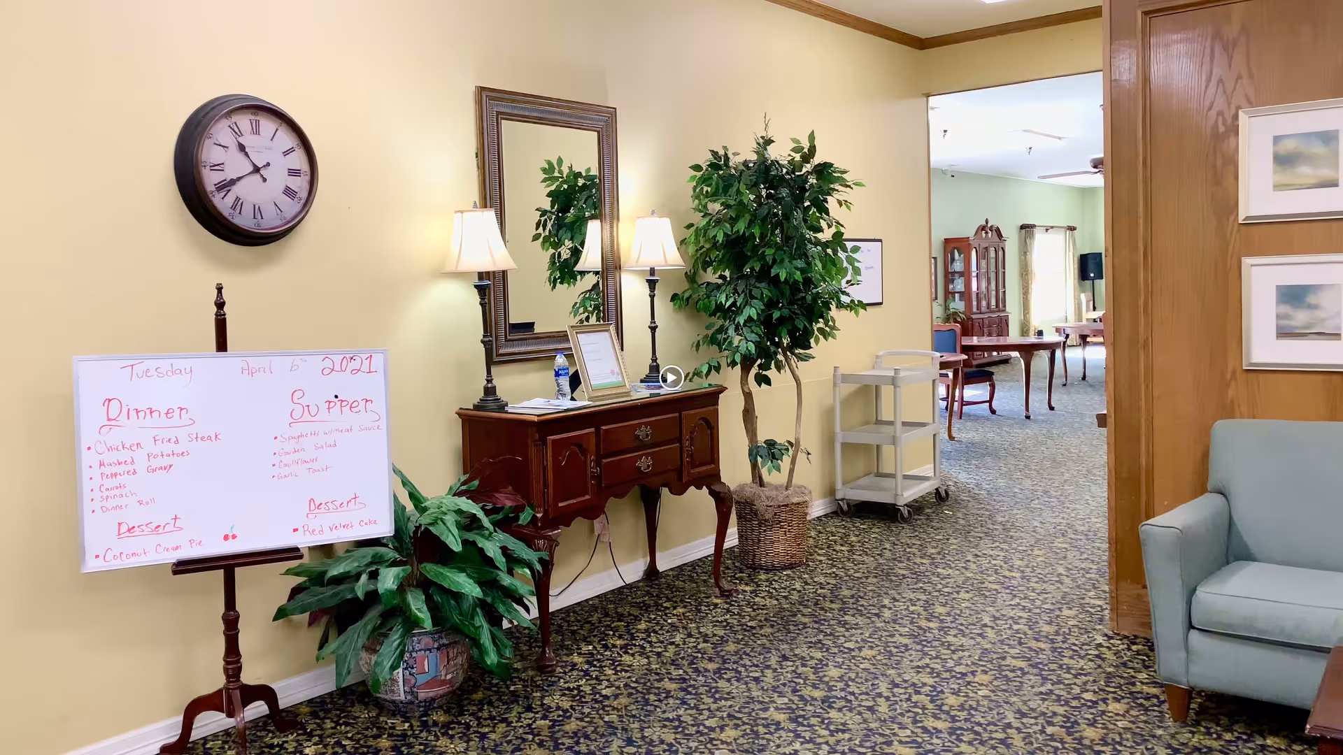 Interior view of a senior living facility hallway with a clock on the wall, a whiteboard displaying dinner and supper menus, a wooden side table with lamps and a mirror above it, potted plants, and a glimpse into a dining area with tables and chairs.
