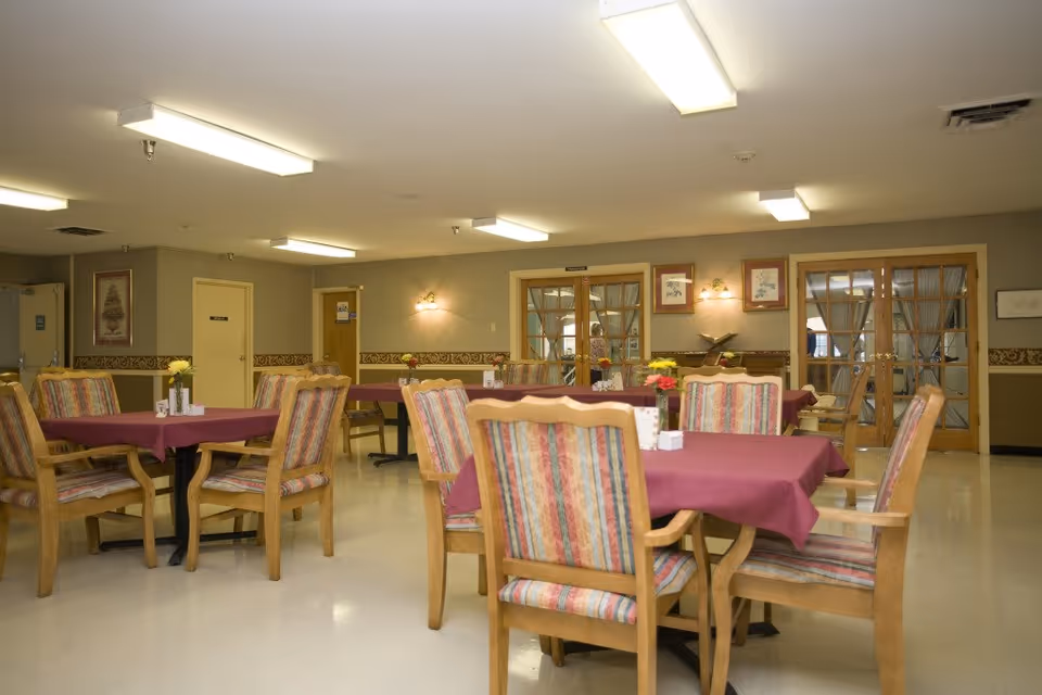Interior view of a dining room with several wooden tables covered with maroon tablecloths and surrounded by cushioned chairs with striped upholstery. The room has beige walls with framed artwork, wall sconces, and fluorescent ceiling lights. There are double glass doors with curtains in the background.