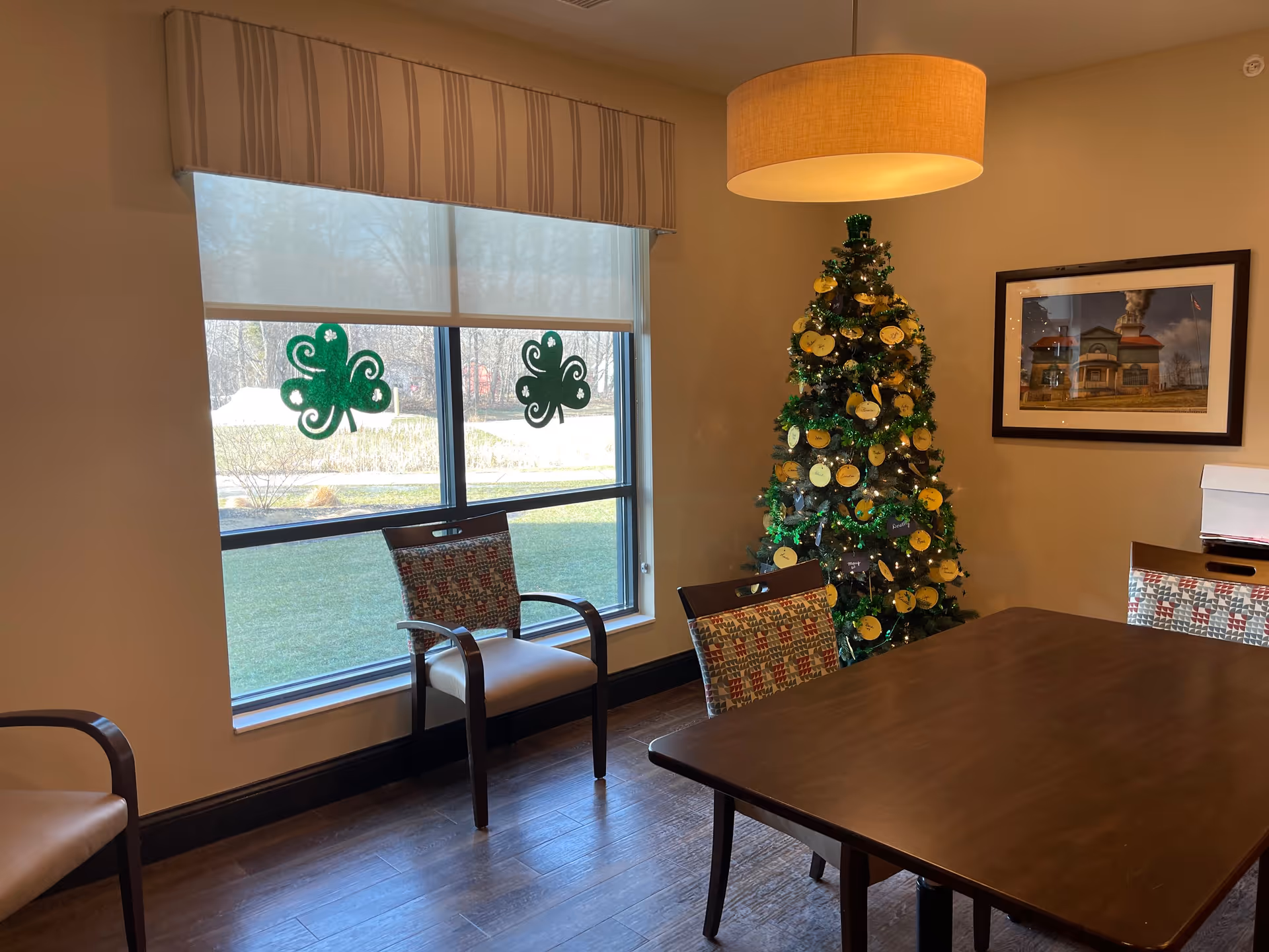 A room with a wooden table and several chairs with patterned cushions. There is a decorated Christmas tree with green and gold ornaments in the corner. Two green shamrock decorations are on the window, which shows a grassy outdoor area. A framed picture hangs on the wall, and a round ceiling light fixture is above the table.