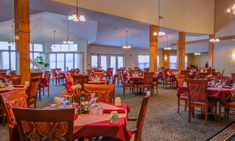 Spacious dining room with many tables covered in red tablecloths and set with glassware and chairs under hanging lights.