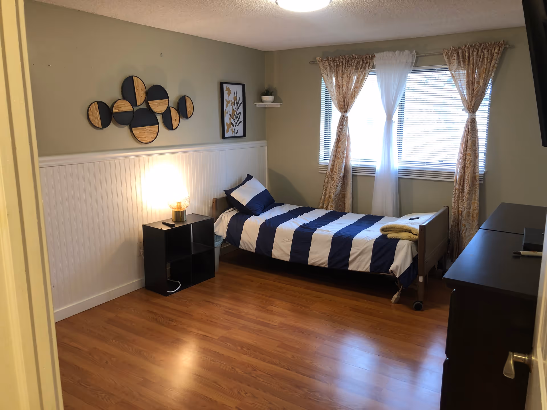 A simple bedroom with a single bed featuring a blue and white striped comforter and pillow. Next to the bed is a black nightstand with a lamp on it. The room has wooden flooring, light green walls with white wainscoting, and a window with patterned curtains and blinds. There is wall art above the bed and a dark dresser on the right side of the room.