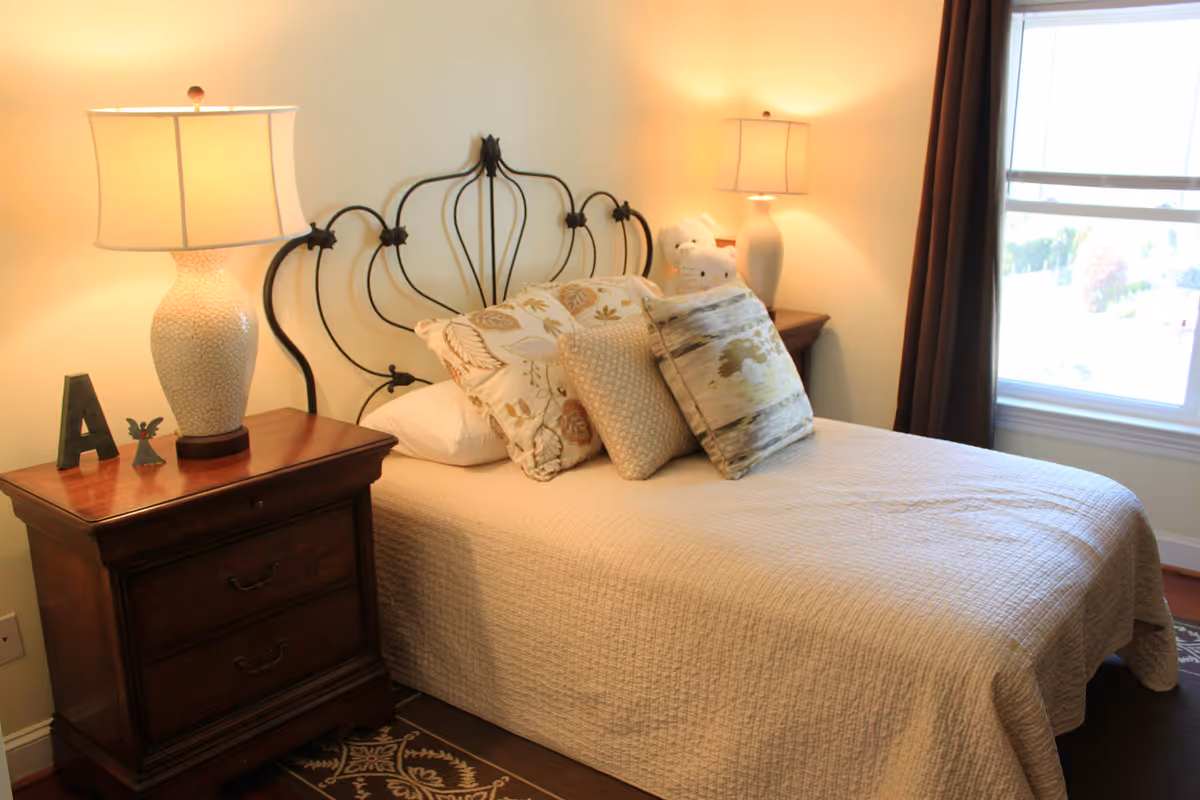 Bedroom in Vienna Manor Assisted Living with a metal headboard, two lamps, and decorative pillows.