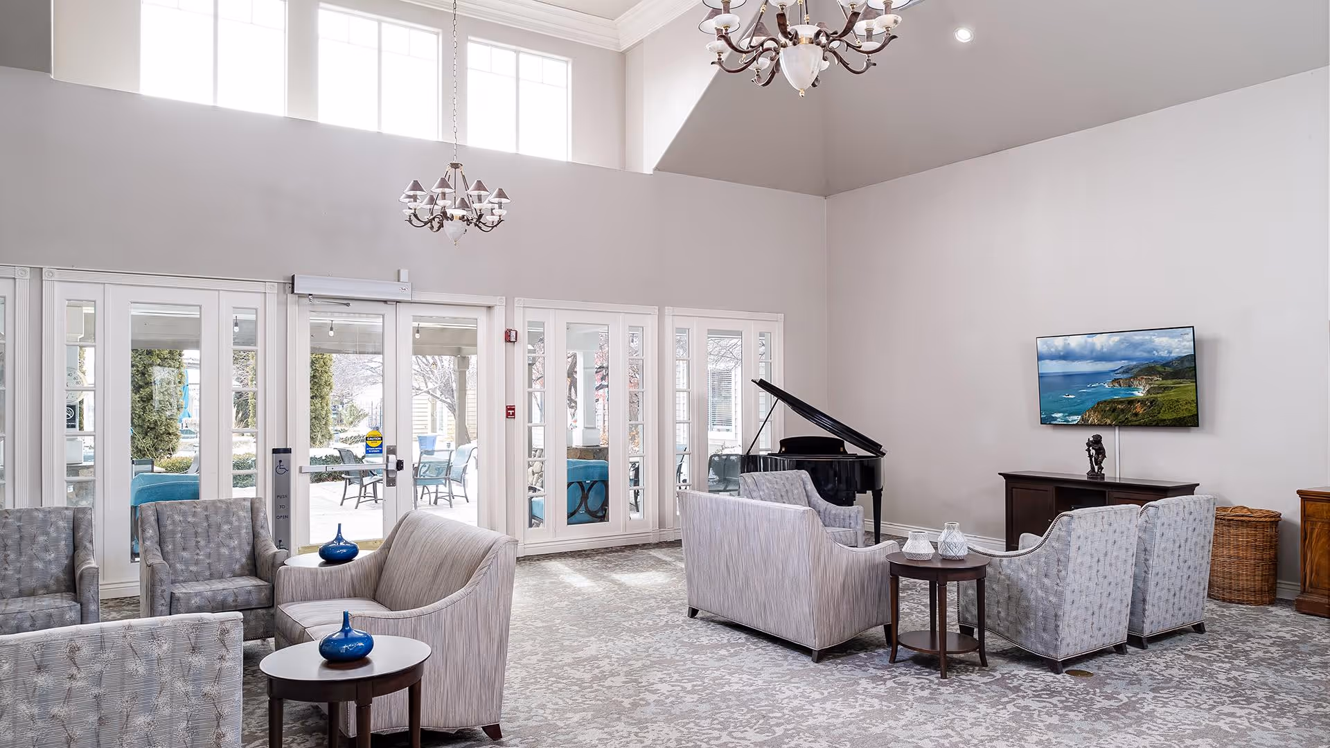 A bright and spacious living room area with high ceilings and large windows. The room features multiple gray upholstered chairs and sofas arranged around small round tables. A black grand piano is positioned near the wall, and a flat-screen TV is mounted above a wooden cabinet. Two chandeliers hang from the ceiling, and glass doors lead to an outdoor patio area with seating.