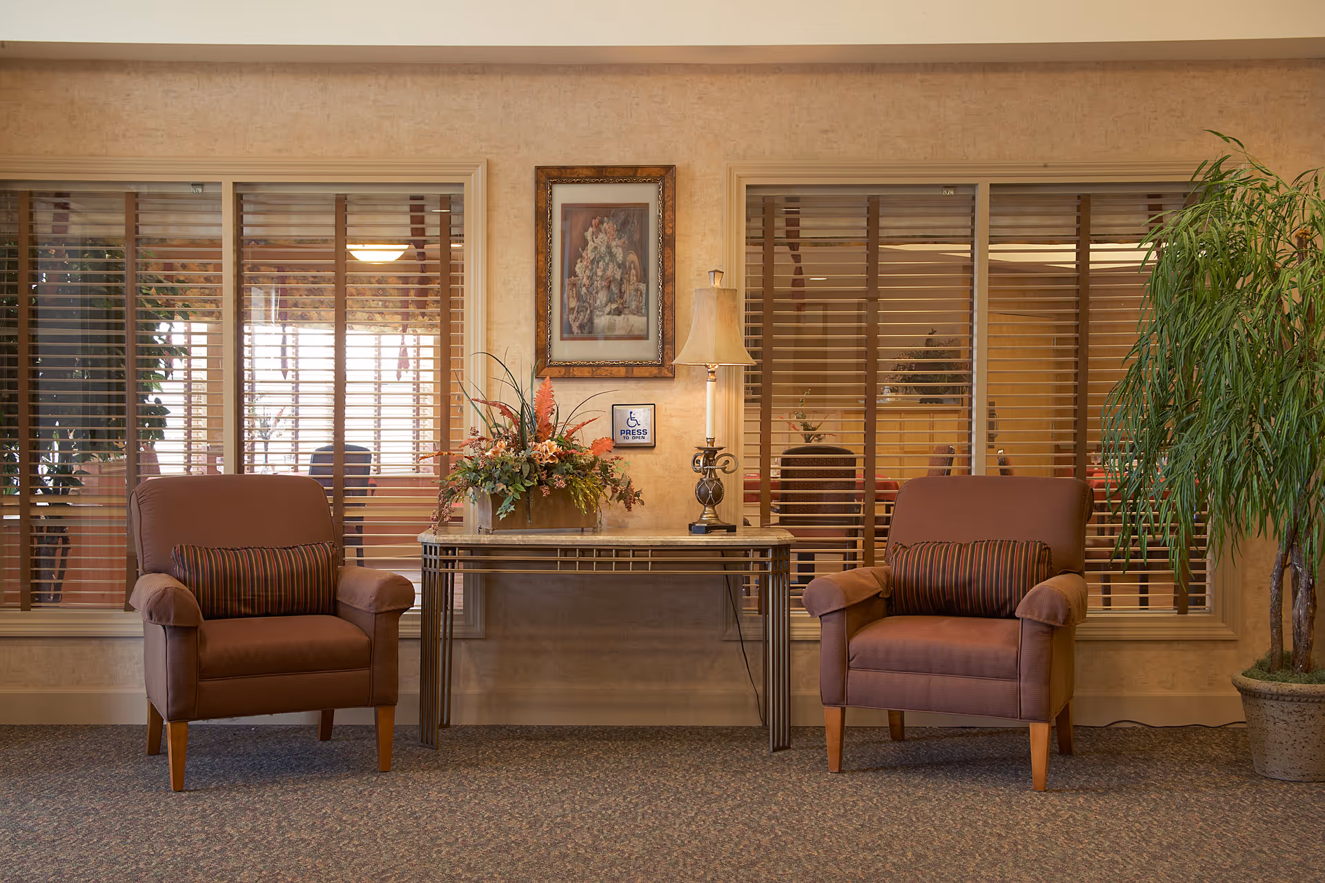 A cozy seating area with two brown armchairs, each with a striped cushion, positioned on either side of a glass-top table. The table holds a decorative floral arrangement and a lamp. Behind the seating area are two large windows with horizontal blinds, and a framed floral painting hangs on the wall between the windows. A potted plant is placed on the right side of the scene.