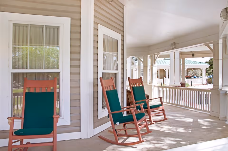 A covered porch area with three wooden rocking chairs featuring green cushions, beige siding walls, white framed windows, and white railings. The porch overlooks a driveway and other parts of the building with green roofs and white columns.