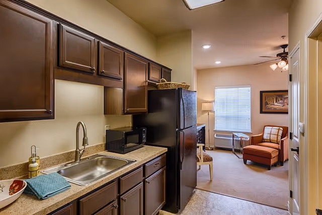 Kitchenette with dark wood cabinets, sink and refrigerator opening into a cozy living area with a chair and window.