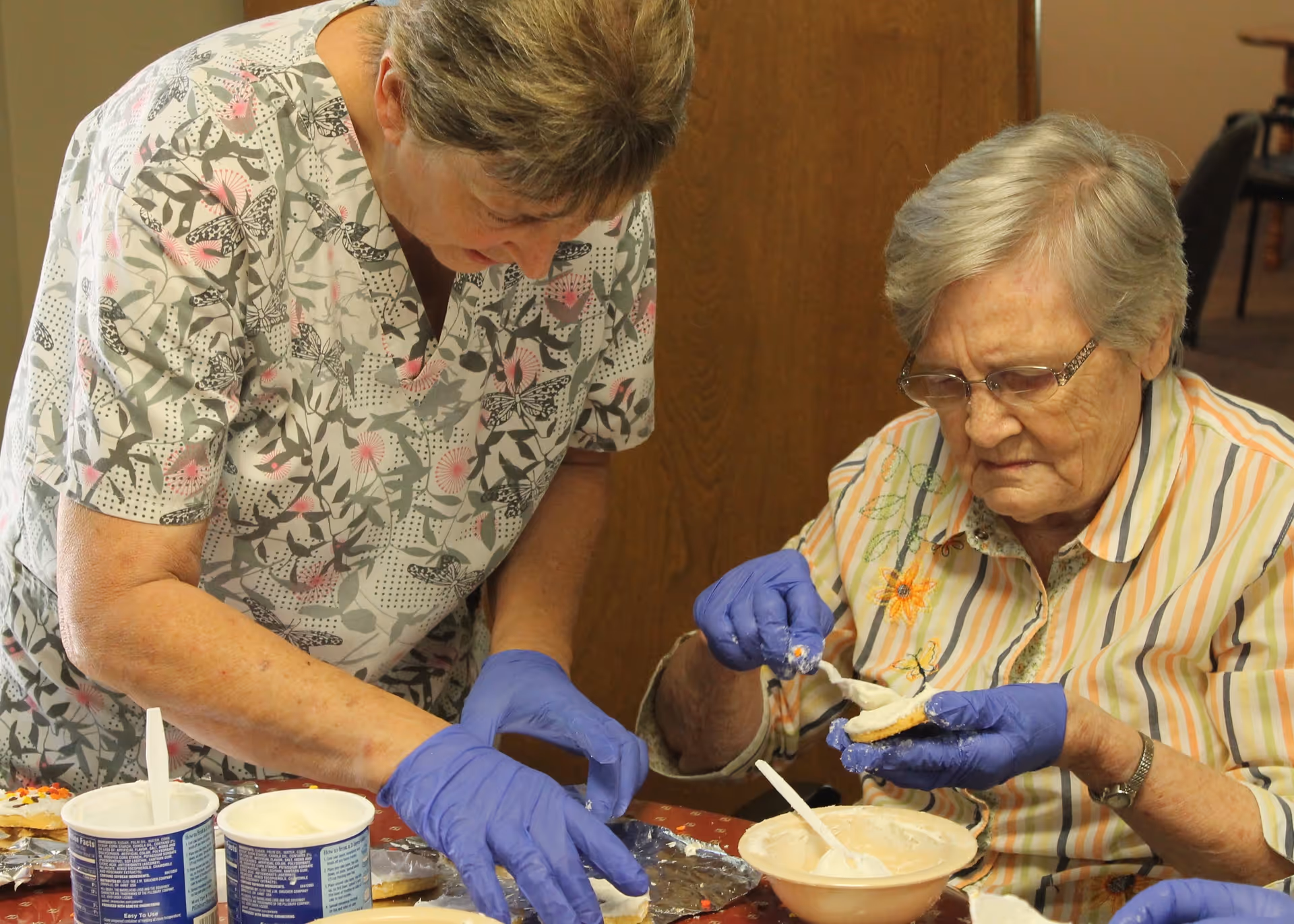 Two elderly women wearing gloves are sitting at a table and decorating cookies with white frosting. One woman is spreading frosting on a cookie while the other is adding sprinkles. Various containers of frosting and sprinkles are on the table.