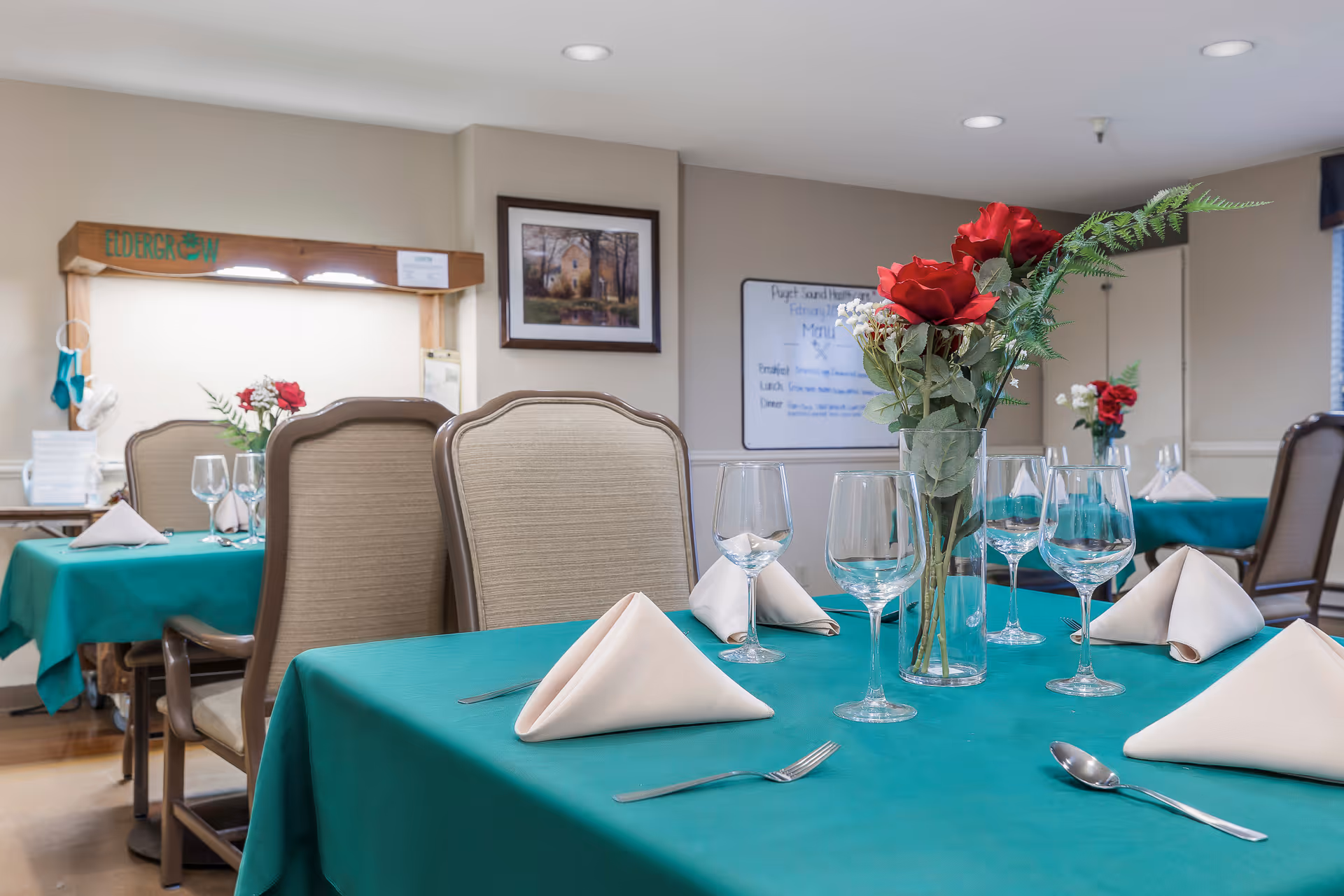 Dining room with tables covered in teal tablecloths, each set with folded white napkins, wine glasses, and silverware. A vase with red roses and greenery is placed in the center of the nearest table. Beige upholstered chairs surround the tables. The room has neutral-colored walls with framed artwork and a whiteboard displaying a menu in the background.