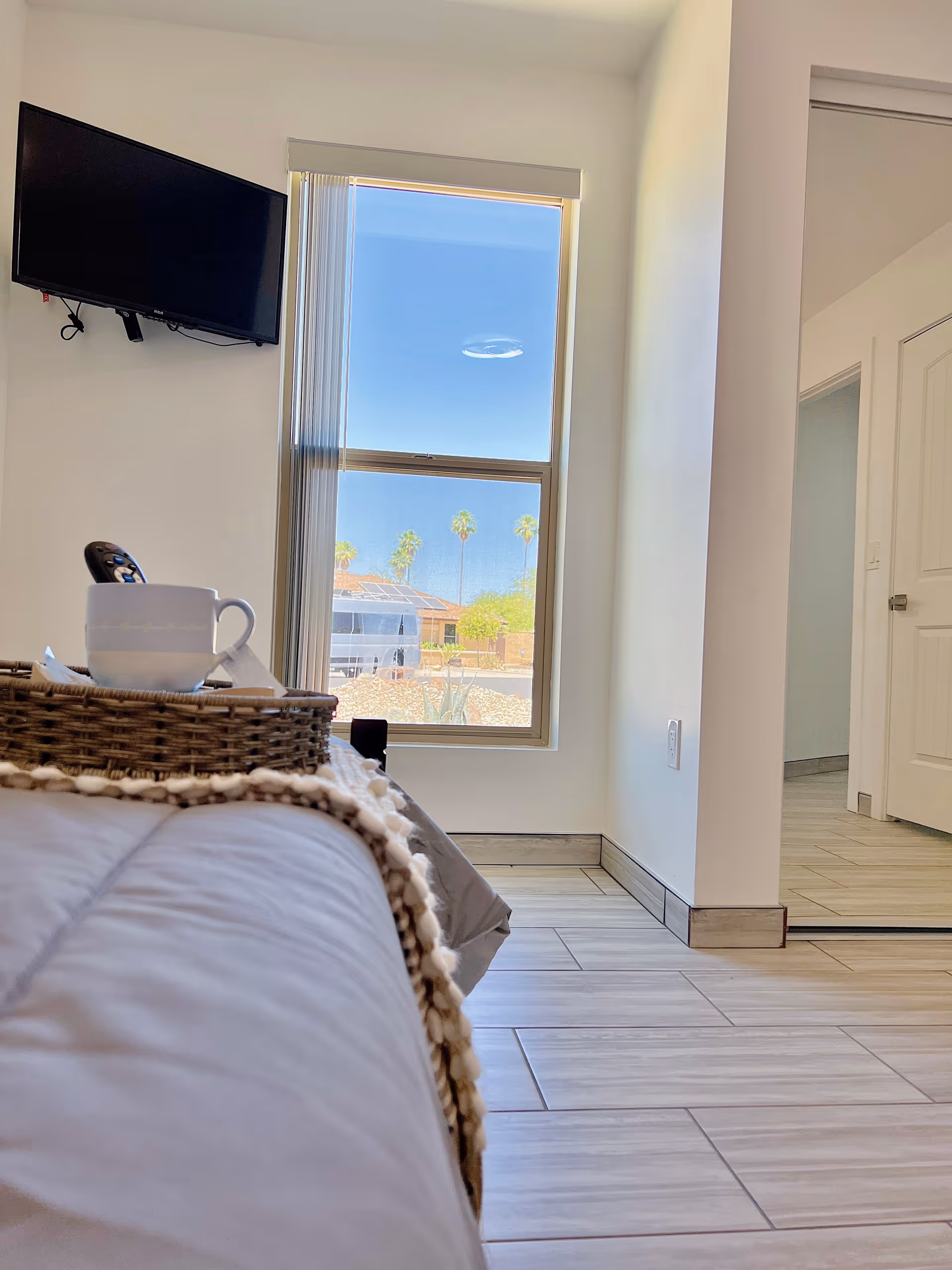 View of a bedroom corner with a bed in the foreground, a wicker tray holding a white cup and remote control on the bed, a window showing a sunny outdoor scene with palm trees, a wall-mounted TV, and a door leading to another room.