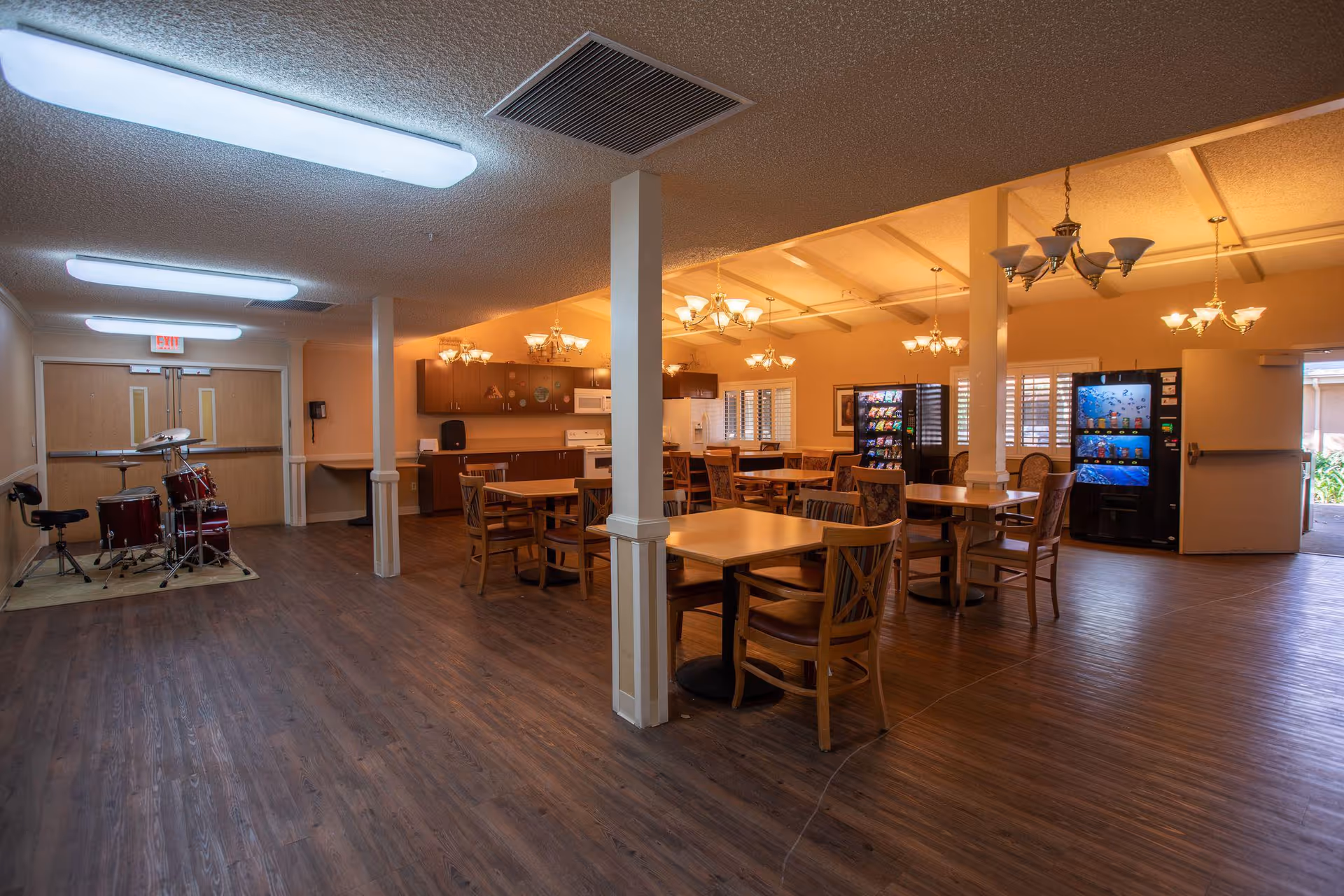 A spacious common area in a senior living facility featuring multiple wooden tables and chairs arranged for dining or social activities. The room has warm lighting with several chandeliers hanging from the ceiling. There is a kitchen area with cabinets, a microwave, and a stove in the background. Two vending machines are visible near an open door leading outside. On the left side of the room, there is a drum set placed on a small rug near double doors. The floor is covered with wood-like flooring.