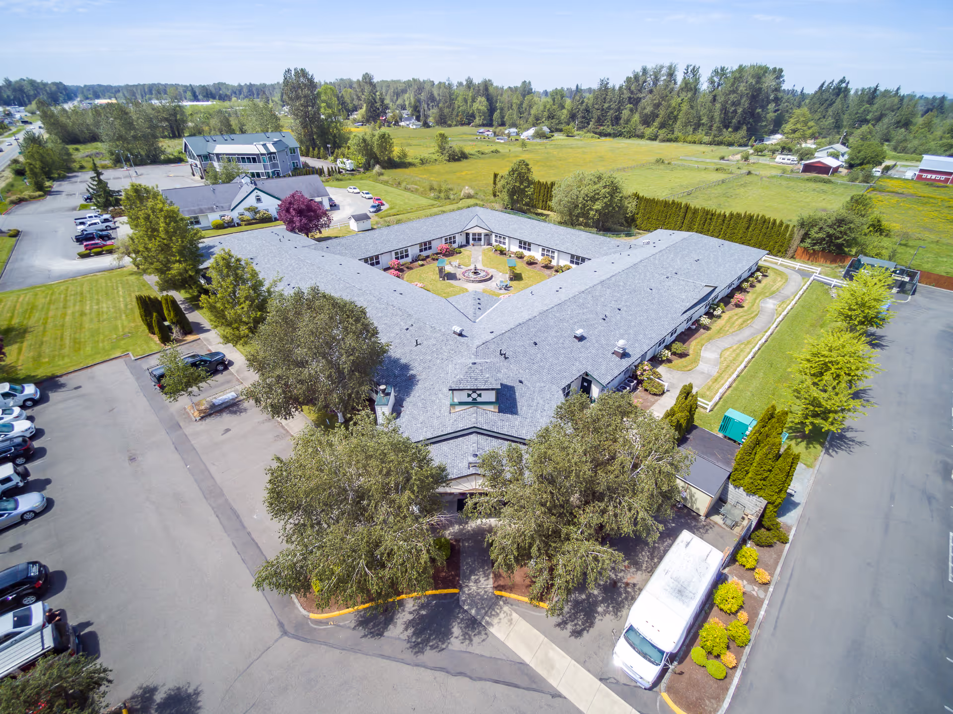 Aerial view of Heritage House Buckley Assisted Living & Memory Care facility showing a large U-shaped building with a central courtyard featuring a fountain and seating areas. The building is surrounded by parking lots, trees, and green fields under a clear sky.