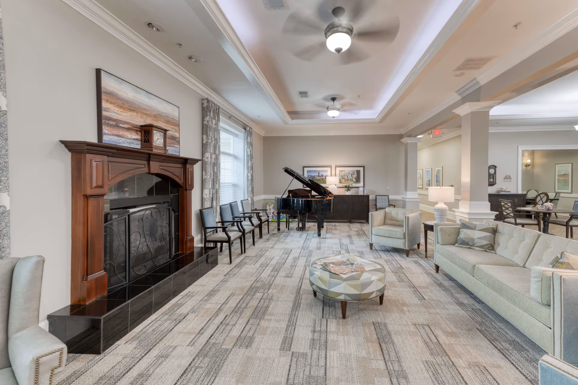 A spacious and well-lit living room area in a senior living facility featuring a wooden fireplace with a clock and artwork above it, several chairs lined up against the wall, a grand piano, a patterned round ottoman, a light green sofa with pillows, and armchairs. The room has ceiling fans, large windows with curtains, and a carpeted floor with a geometric pattern.
