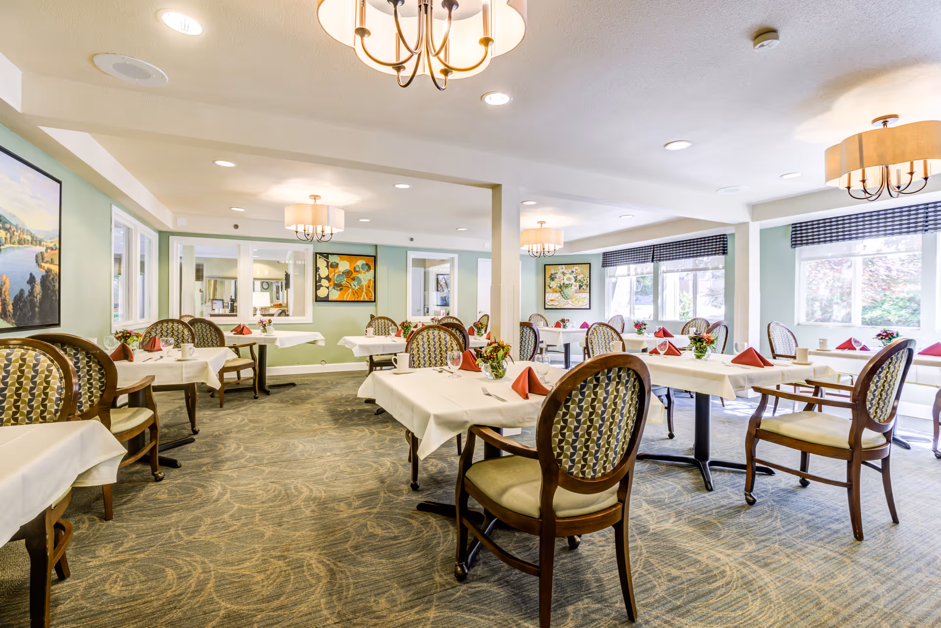 Dining room with multiple tables set with white linens, folded red napkins, floral centerpieces, and patterned chairs.