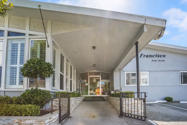 Covered entrance and ramp flanked by low stone planters leading to the glass doors of the San Francisco Post Acute building.
