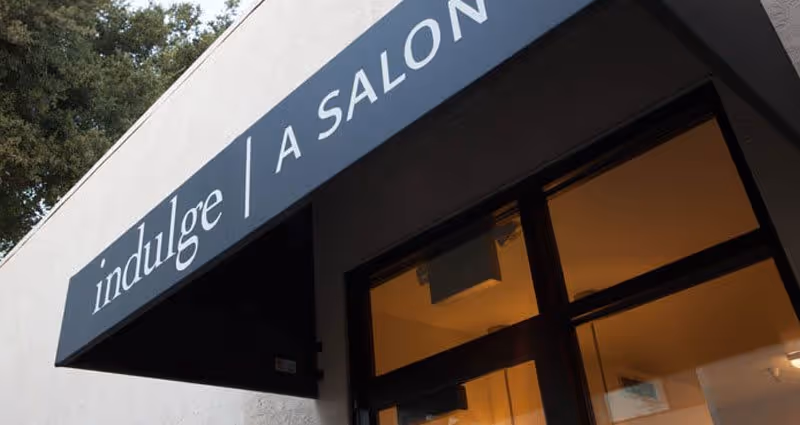 Exterior view of a salon entrance with a black awning displaying the text 'indulge | A SALON'. The building is light-colored with a glass door and window showing warm indoor lighting.