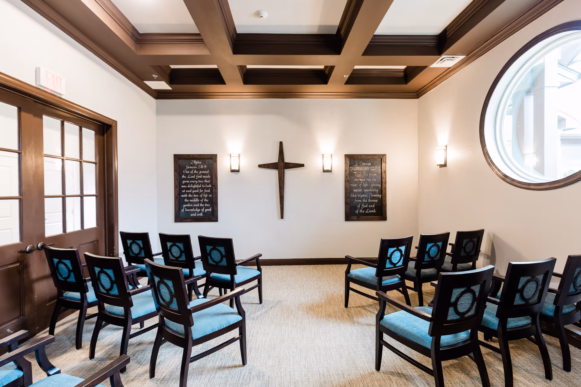 A small chapel or meditation room with wooden chairs arranged in rows facing a wall with a wooden cross mounted between two framed scripture plaques. The room has a coffered ceiling with wooden beams, beige carpet, and a round window on the right side letting in natural light.