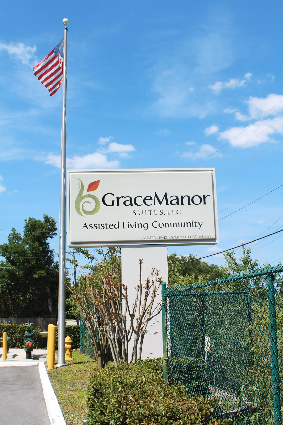 Outdoor sign for Grace Manor Suites assisted living community beside a flagpole under a blue sky.