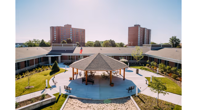 Outdoor courtyard area at Dunklau Gardens featuring a central gazebo with seating underneath, surrounded by paved walkways, landscaped greenery, and a sandpit in the foreground. The courtyard is enclosed by a single-story building, with taller residential buildings visible in the background under a clear sky.
