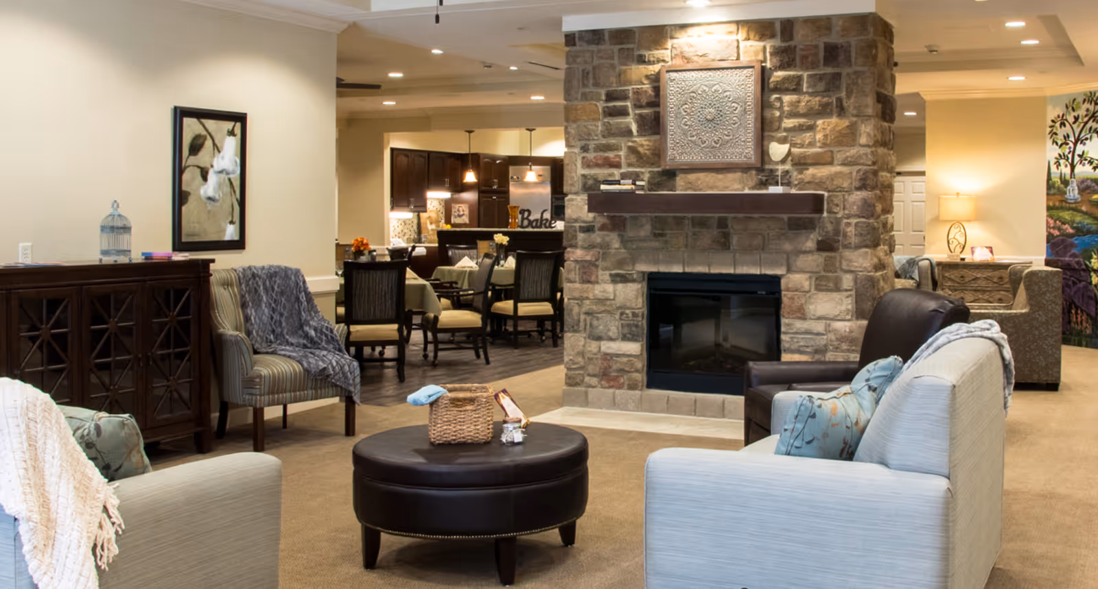 Spacious senior living common area with sofas and armchairs arranged around a central ottoman and a stone fireplace, with a dining area visible in the background.