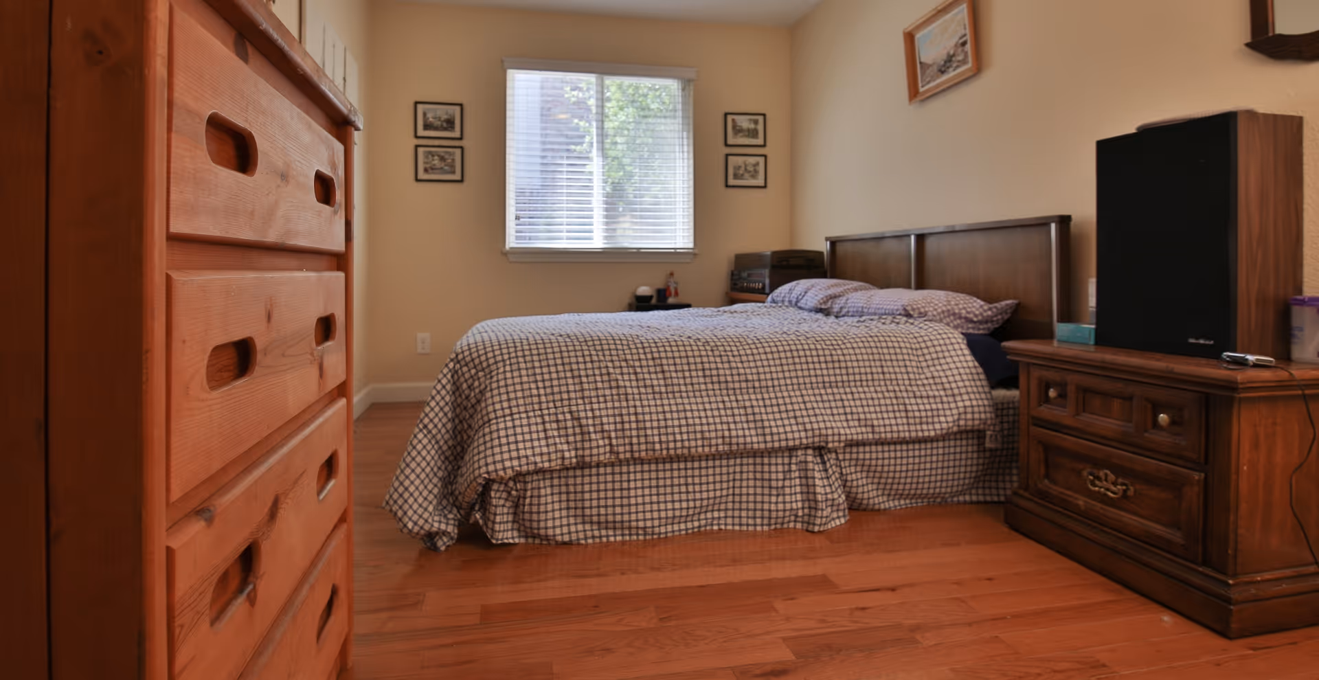 A bedroom with a wooden bed frame and a bed covered with a checkered bedspread. There is a wooden dresser on the left side and a wooden nightstand with a large speaker on the right side. A window with blinds is centered on the far wall, flanked by four small framed pictures. The floor is hardwood, and the walls are painted beige.
