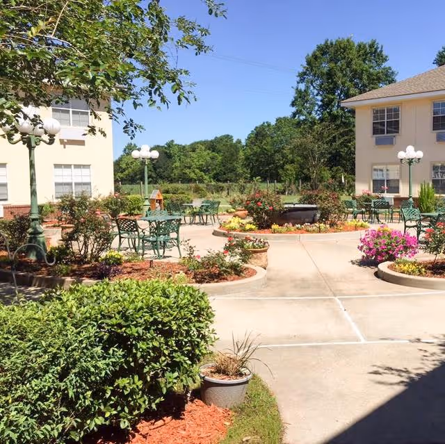 Outdoor courtyard area of an assisted living facility with paved walkways, green metal tables and chairs, flower beds with colorful flowers, bushes, and lamp posts. Two beige buildings with windows flank the courtyard, and trees and greenery are visible in the background under a clear blue sky.