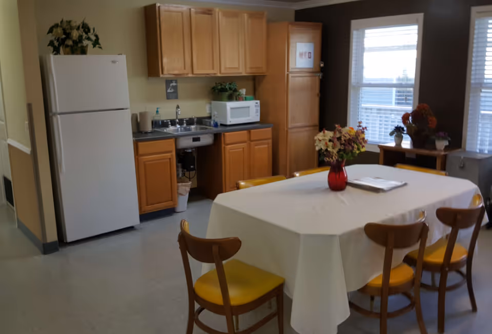 A small kitchen and dining area with wooden cabinets, a white refrigerator, a microwave on the counter, a sink, and a table covered with a white tablecloth surrounded by six wooden chairs with yellow cushions. There are flowers in a red vase on the table and additional plants near the windows.