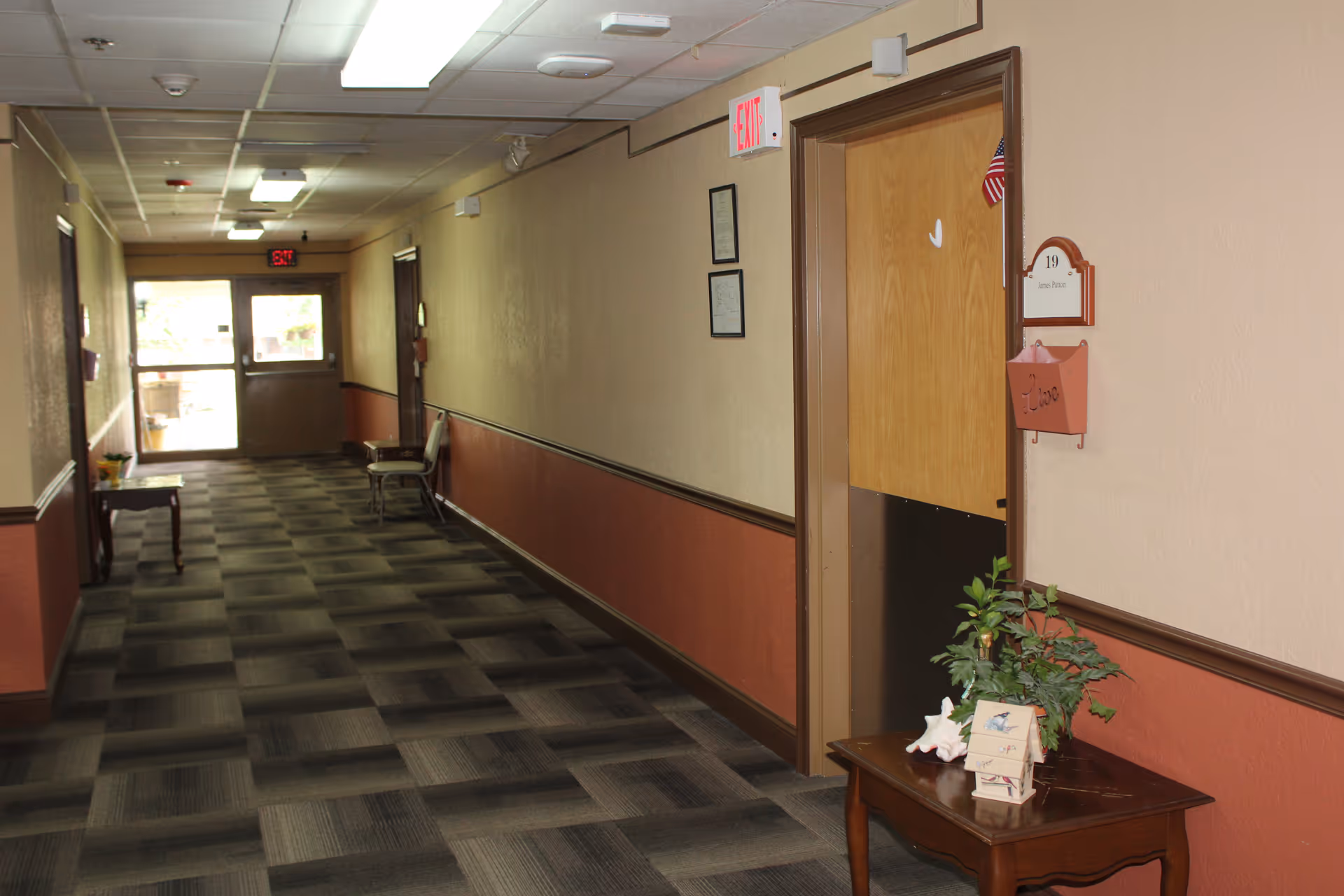 A long indoor hallway with patterned carpet and beige walls with a brown lower half. Several doors line the hallway, one door labeled 19 with a small American flag and a pink mailbox attached. A small wooden table with a plant, a shell, and a decorative item is placed near the door. There are chairs and small tables along the hallway, and exit signs are visible at both ends.