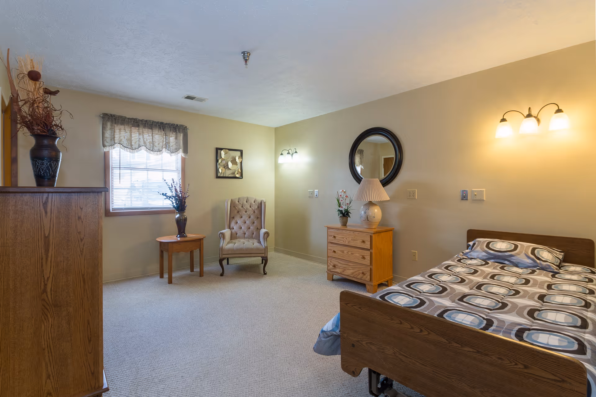 A cozy bedroom in a senior living facility featuring a single bed with patterned bedding, a wooden dresser with a lamp and a small plant, a cushioned armchair next to a small round table with a vase of flowers, a window with a valance, and soft yellow walls with wall-mounted lights.
