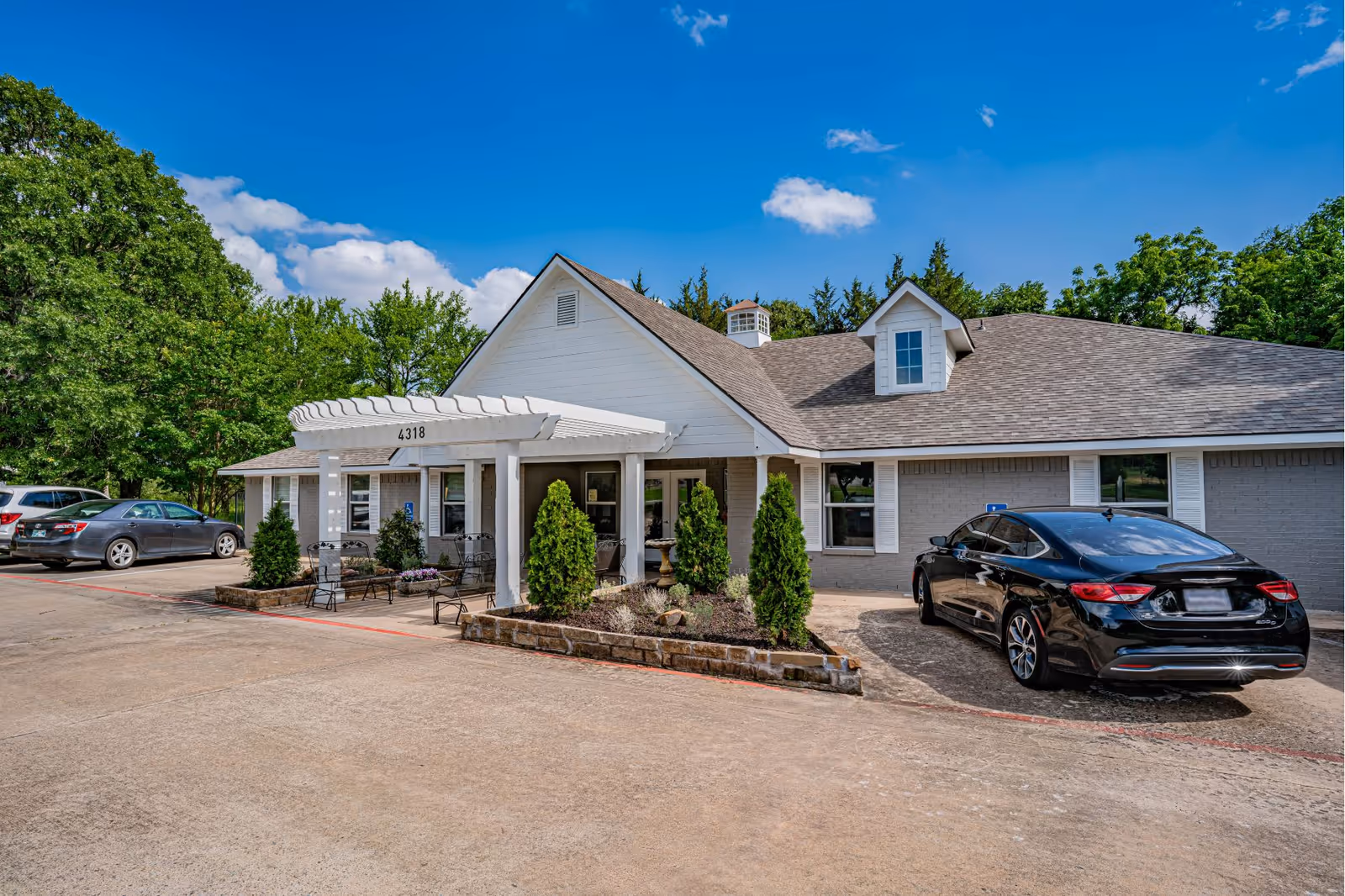 Exterior view of a single-story assisted living facility building with a white pergola entrance labeled 4318. Several cars are parked in front, and there are small landscaped areas with shrubs and trees around the building. The sky is clear with a few clouds.
