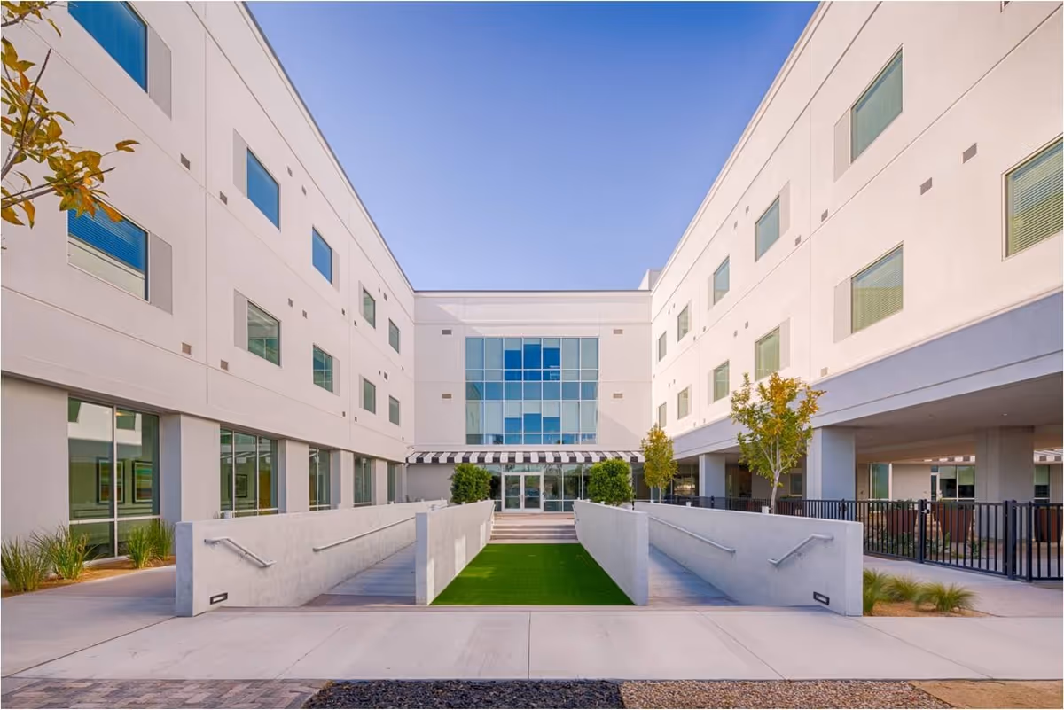 Exterior view of a modern three-story building with large windows and a central entrance. There is a ramp with handrails leading up to the entrance, flanked by small trees and landscaping. The sky is clear and blue.