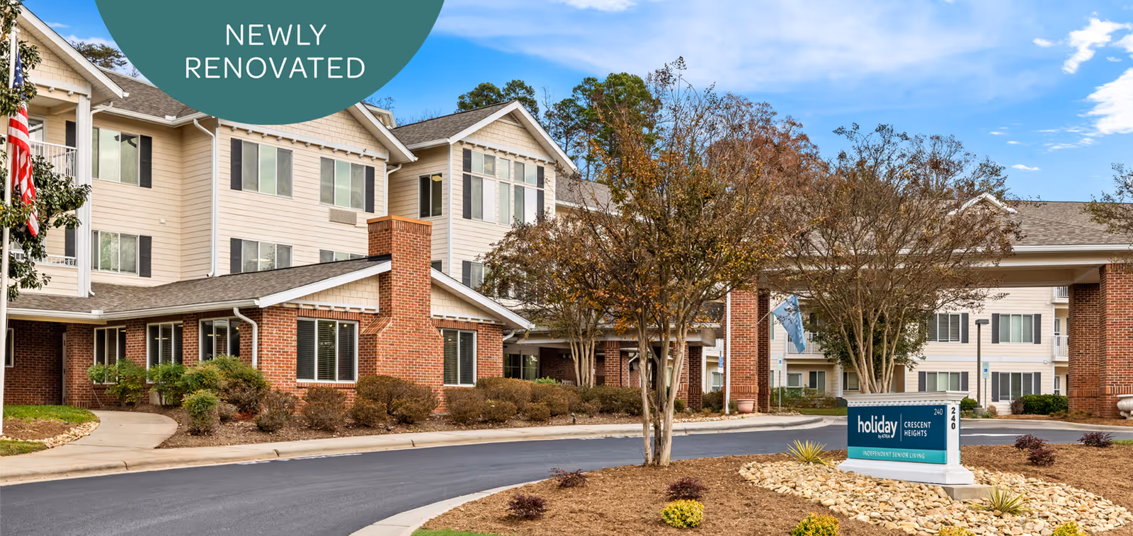 Exterior view of Holiday Crescent Heights senior living facility showing a newly renovated building with beige siding and brick accents. The entrance features a covered drop-off area, landscaped grounds with trees and shrubs, and a sign displaying the facility's name.