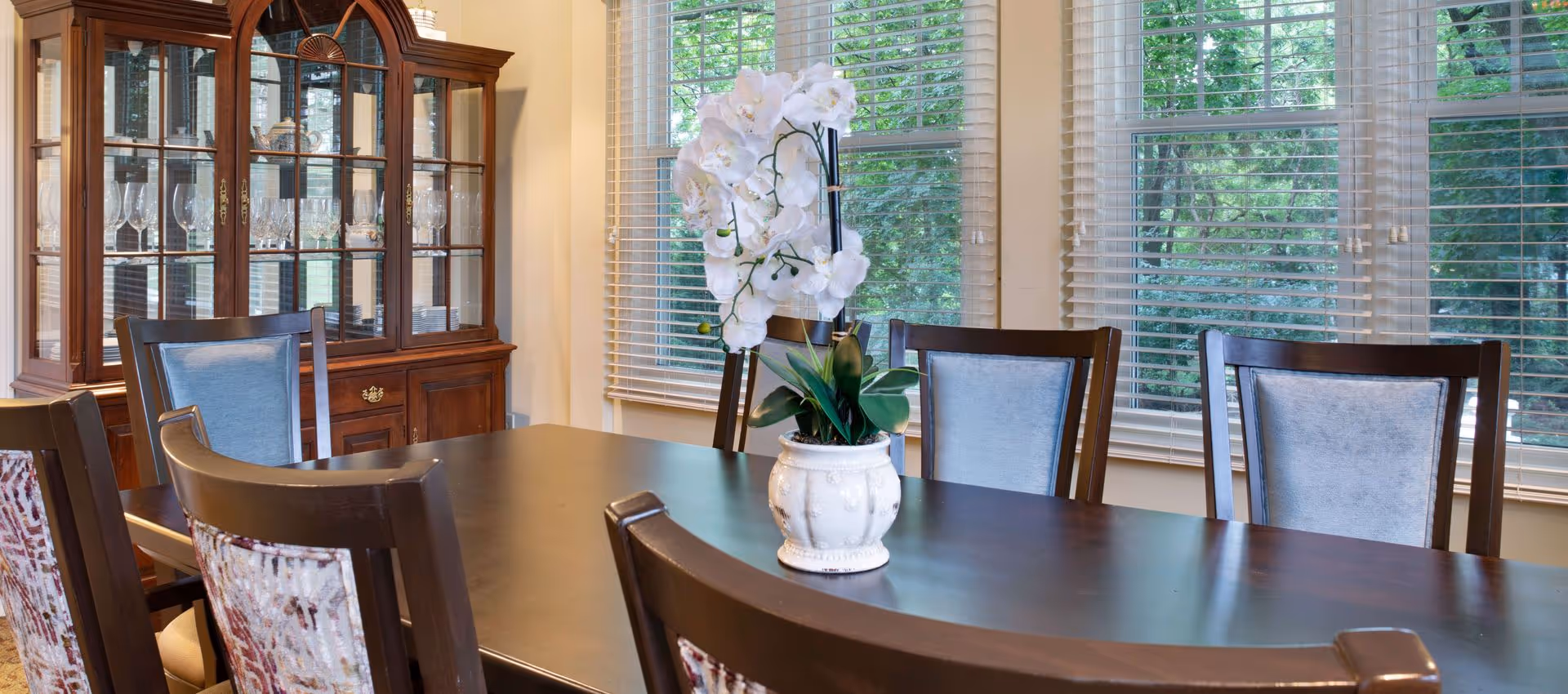 A bright dining room with a dark wood table and chairs, a white orchid centerpiece, a china cabinet, and large windows with blinds.