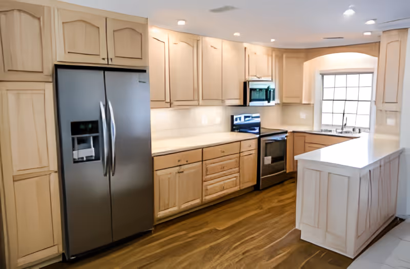 A modern kitchen with light wood cabinets, a stainless steel refrigerator with a water dispenser, a built-in microwave above a stainless steel oven, a white countertop, and a window above the sink allowing natural light to enter.