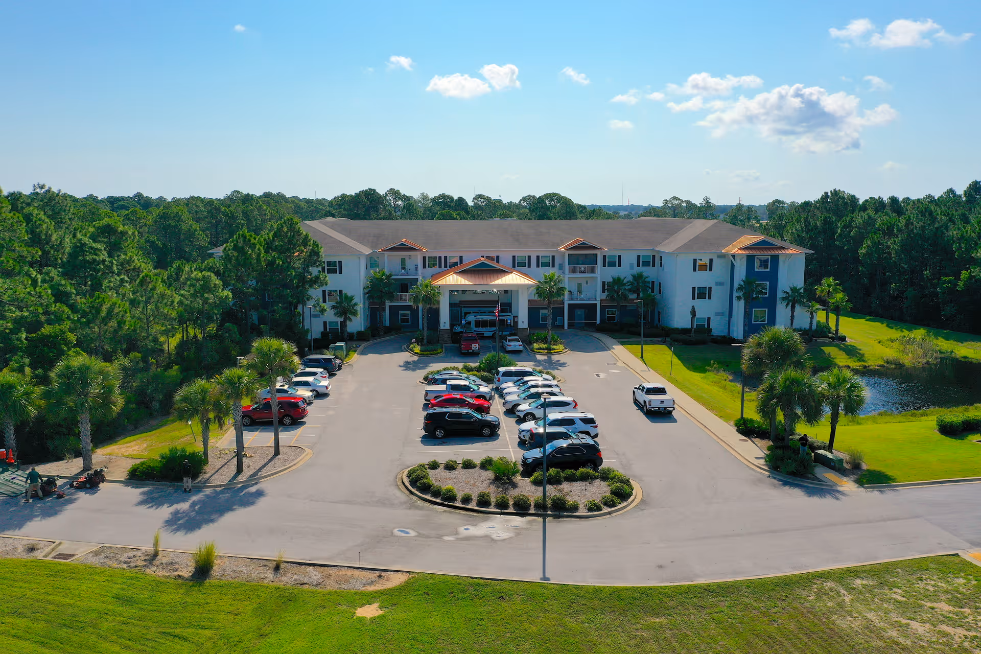 Front view of a three-story senior living building with a parking lot, palm trees, and surrounding greenery under a blue sky.