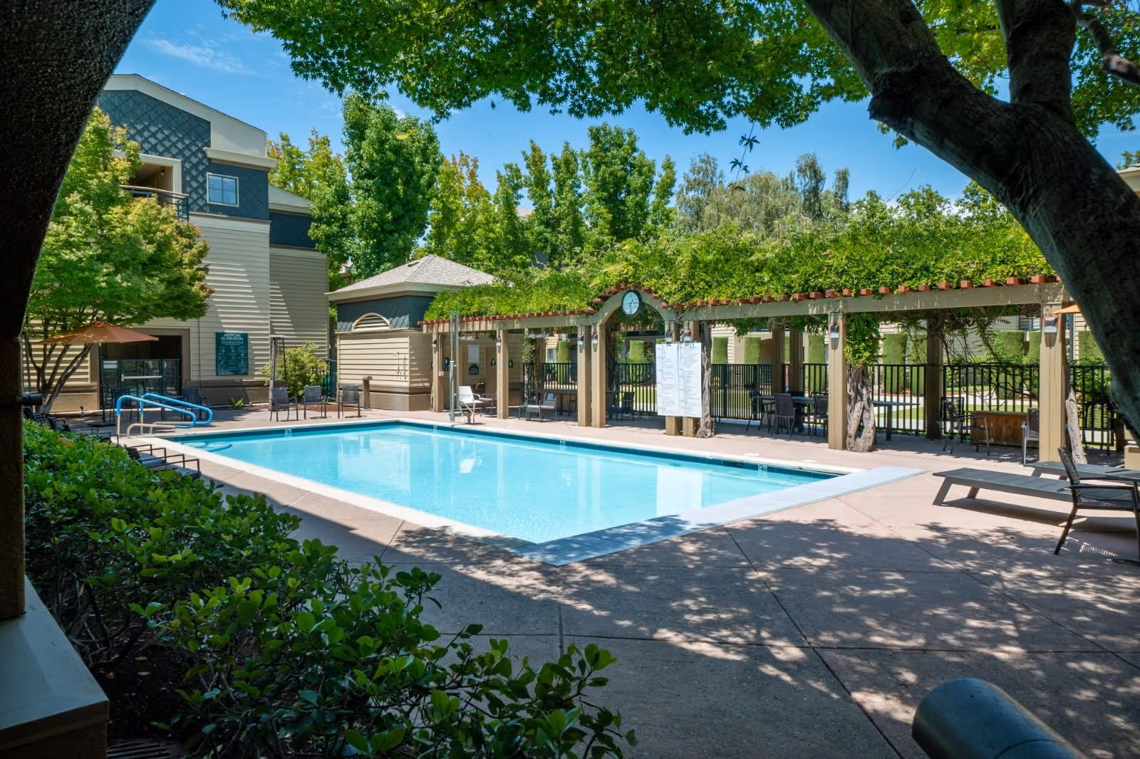 Outdoor swimming pool area at The Terraces of Los Gatos with clear blue water, surrounded by a concrete deck, shaded pergola with greenery on top, seating areas with chairs and tables, and trees providing additional shade.