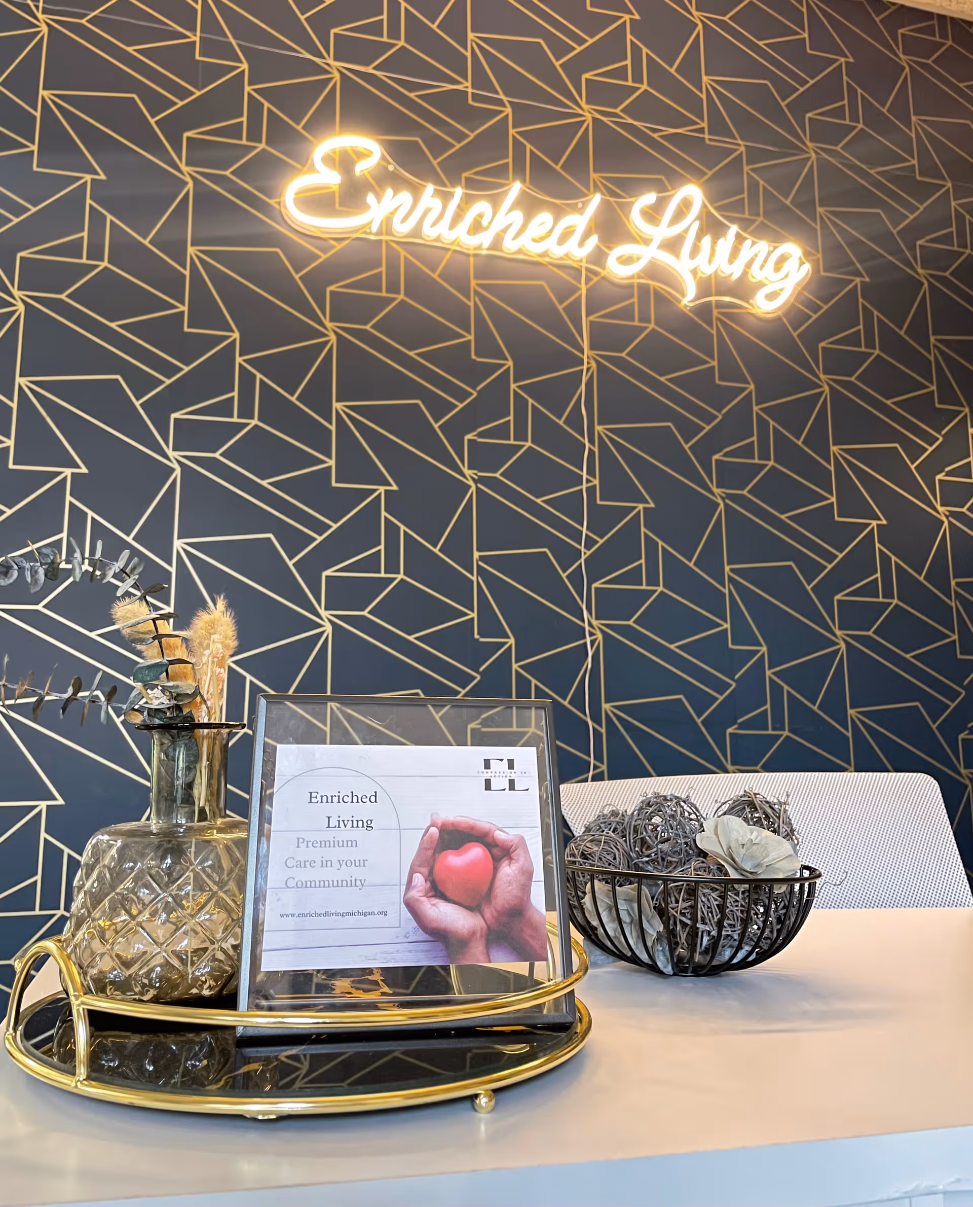 A modern reception or office desk with a decorative glass vase holding dried plants, a framed sign that reads 'Enriched Living Premium Care in your Community', and a black wire bowl filled with decorative wicker balls and dried flowers. Behind the desk is a dark wall with a gold geometric pattern and a glowing neon sign that says 'Enriched Living'.