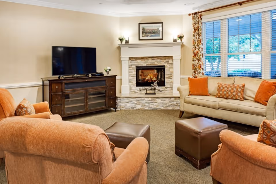 A cozy living room with beige walls and carpet, featuring a stone fireplace with a fire burning, a flat-screen TV on a dark wooden stand, a beige sofa with orange and patterned cushions, two orange armchairs, two brown leather ottomans, and a window with floral curtains showing greenery outside.