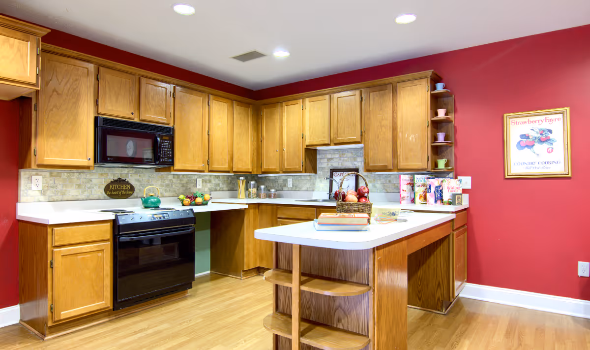 A bright kitchen with wooden cabinets and a white countertop island. The walls are painted red, and there is a black microwave and oven. Various kitchen items, including a basket of apples, cereal boxes, and colorful cups on a small shelf, are displayed on the counters. A framed picture hangs on the wall.
