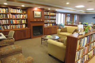 Cozy living room area in a senior living facility with bookshelves filled with books, a fireplace, comfortable armchairs and sofas, a glass coffee table with a decorative centerpiece, and large windows letting in natural light.