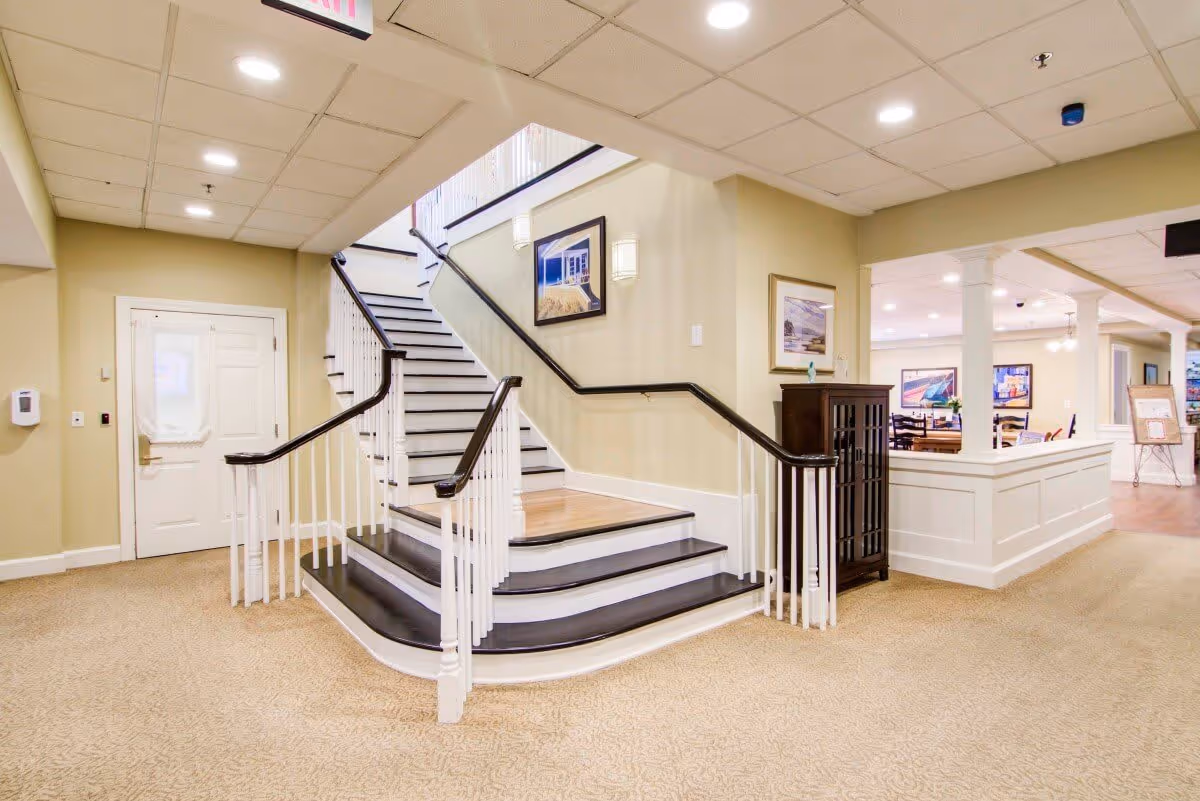 Interior view of a senior living facility showing a carpeted area with a staircase featuring dark wooden handrails and white balusters. The walls are painted light beige with framed pictures hanging. To the right, there is a partial view of a common area with tables and chairs, illuminated by ceiling lights. A white door is visible on the left side of the image.