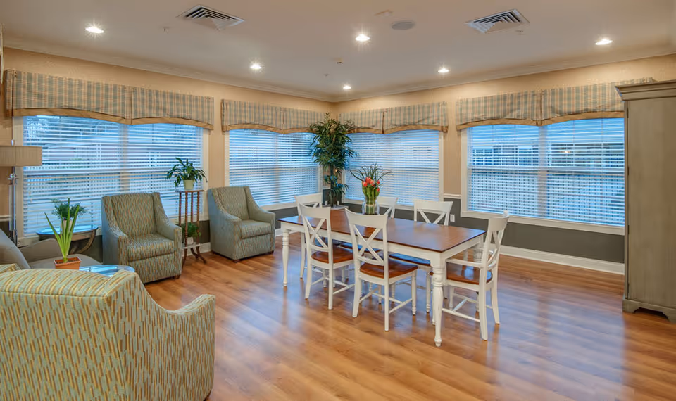 A bright and spacious common area in a senior living facility with large windows covered by plaid valances. The room features a wooden dining table with six white chairs, several upholstered armchairs, potted plants, and wood flooring.