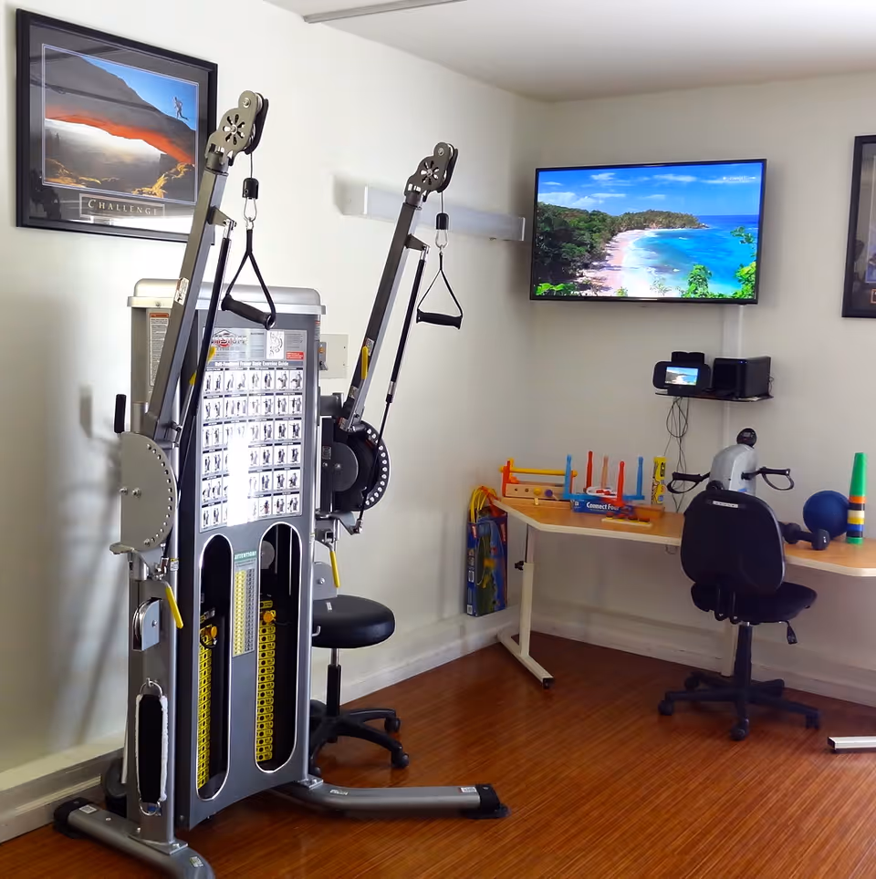 Interior therapy/fitness room with a multi-station exercise machine, chairs, a small activity table with toys, and a wall-mounted TV showing a beach.
