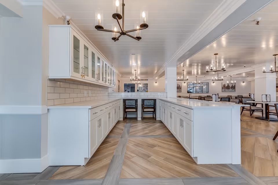 A spacious, modern dining area in Serra Highlands Senior Living featuring white cabinetry with glass doors, a long counter with drawers, and multiple chandeliers hanging from a white ceiling. The room has wood-patterned flooring and several tables and chairs arranged for dining in the background.