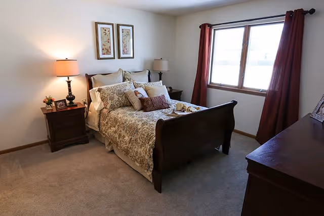 A cozy bedroom with a wooden bed frame, floral bedding, multiple pillows, two bedside tables with lamps, framed artwork on the wall, a large window with red curtains, and a wooden dresser.