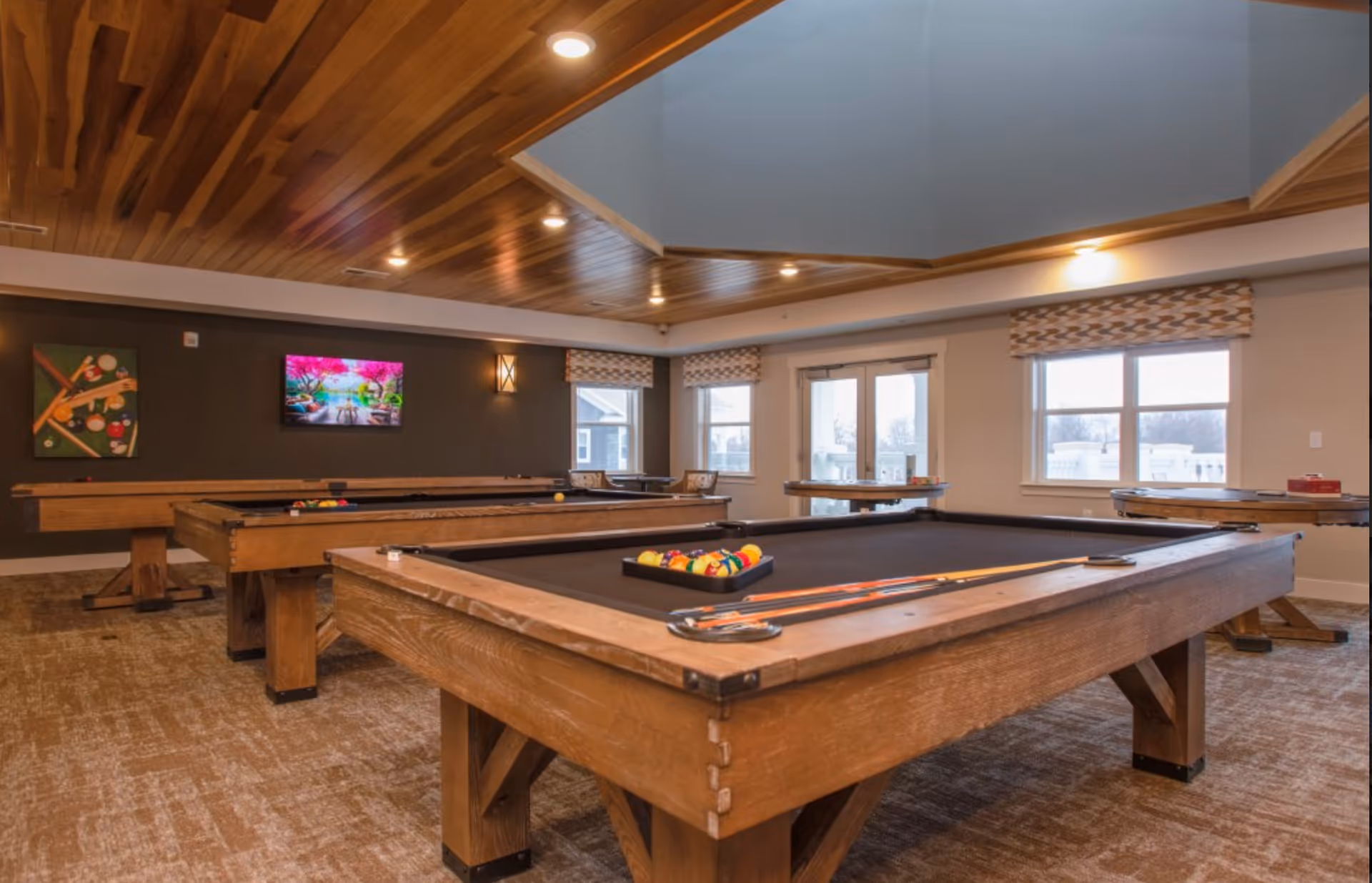 A spacious game room with multiple wooden pool tables, a shuffleboard table, and a wall-mounted TV displaying a colorful scene. The room features a wooden ceiling with recessed lighting, large windows with patterned valances, and carpeted flooring.