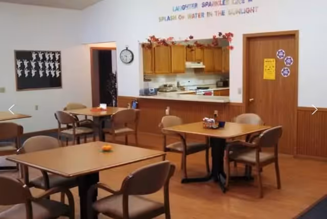 A dining area with several wooden tables and chairs arranged neatly. In the background, there is a kitchen area with wooden cabinets, a stove, and a counter. The walls have decorations including a clock, a bulletin board with cut-out figures, and colorful text that reads 'Laughter Sparkles Like A Splash Of Water In The Sunlight'.