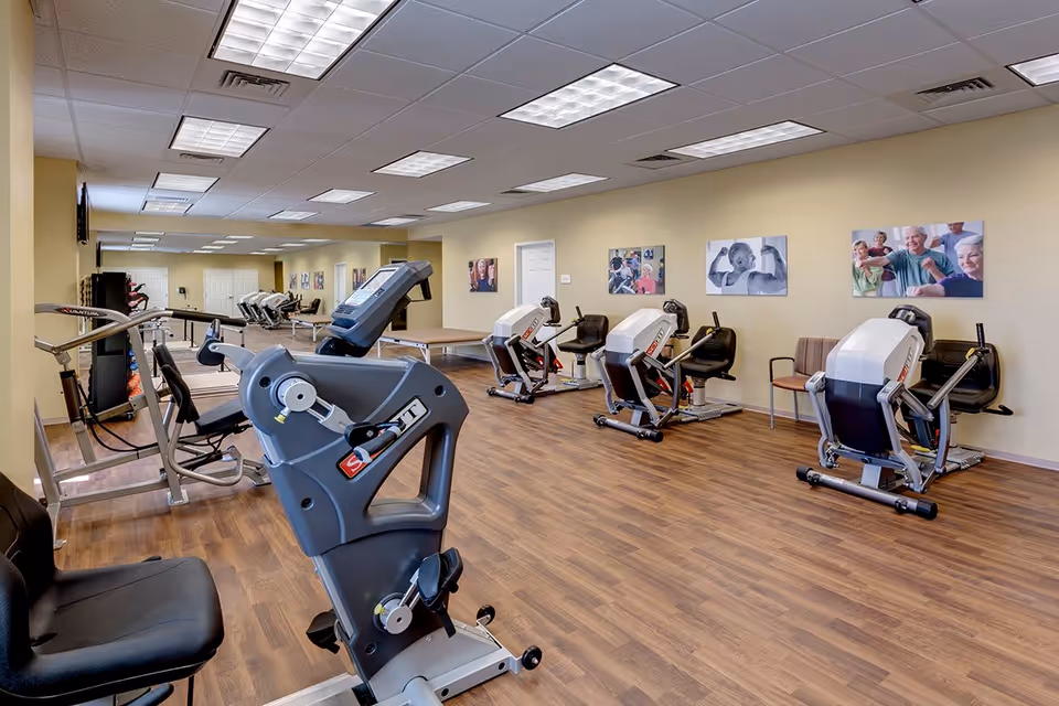 A senior fitness room with various exercise equipment including stationary bikes and resistance machines. The room has wood flooring, beige walls decorated with photos of seniors exercising, and a ceiling with fluorescent lights.