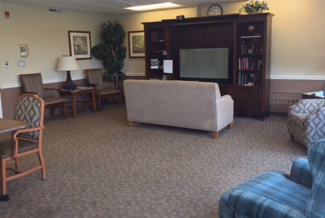 A cozy living room area in a senior living facility with a beige couch facing a large wooden entertainment center with a TV. The room also has several chairs, a side table with a lamp, framed artwork on the walls, a large potted plant, and a carpeted floor.