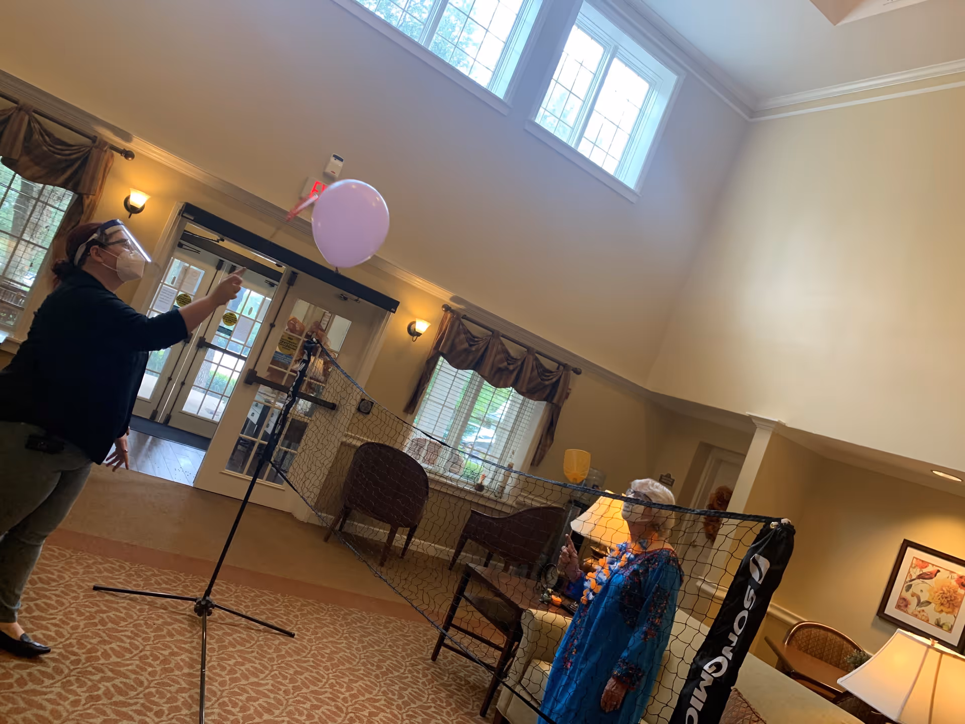 Two people in a senior living community common room play a balloon game over a net, one wearing a face shield.