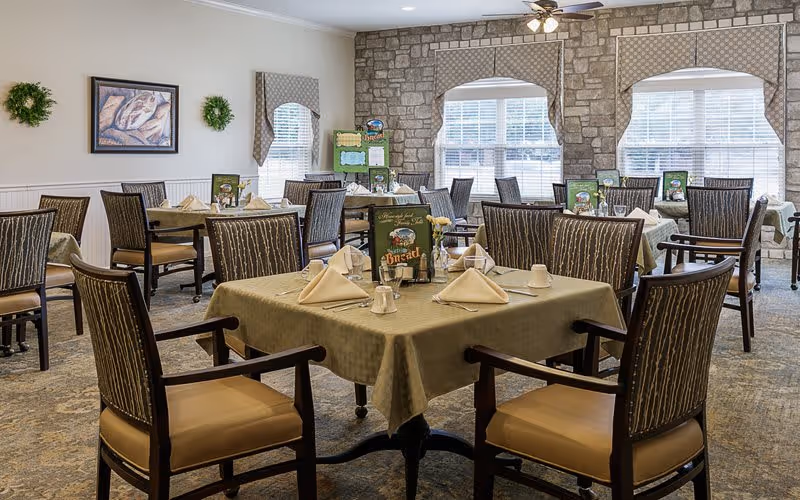 Bright dining room with multiple tables set with napkins and tableware and upholstered chairs in front of a stone accent wall and large windows.